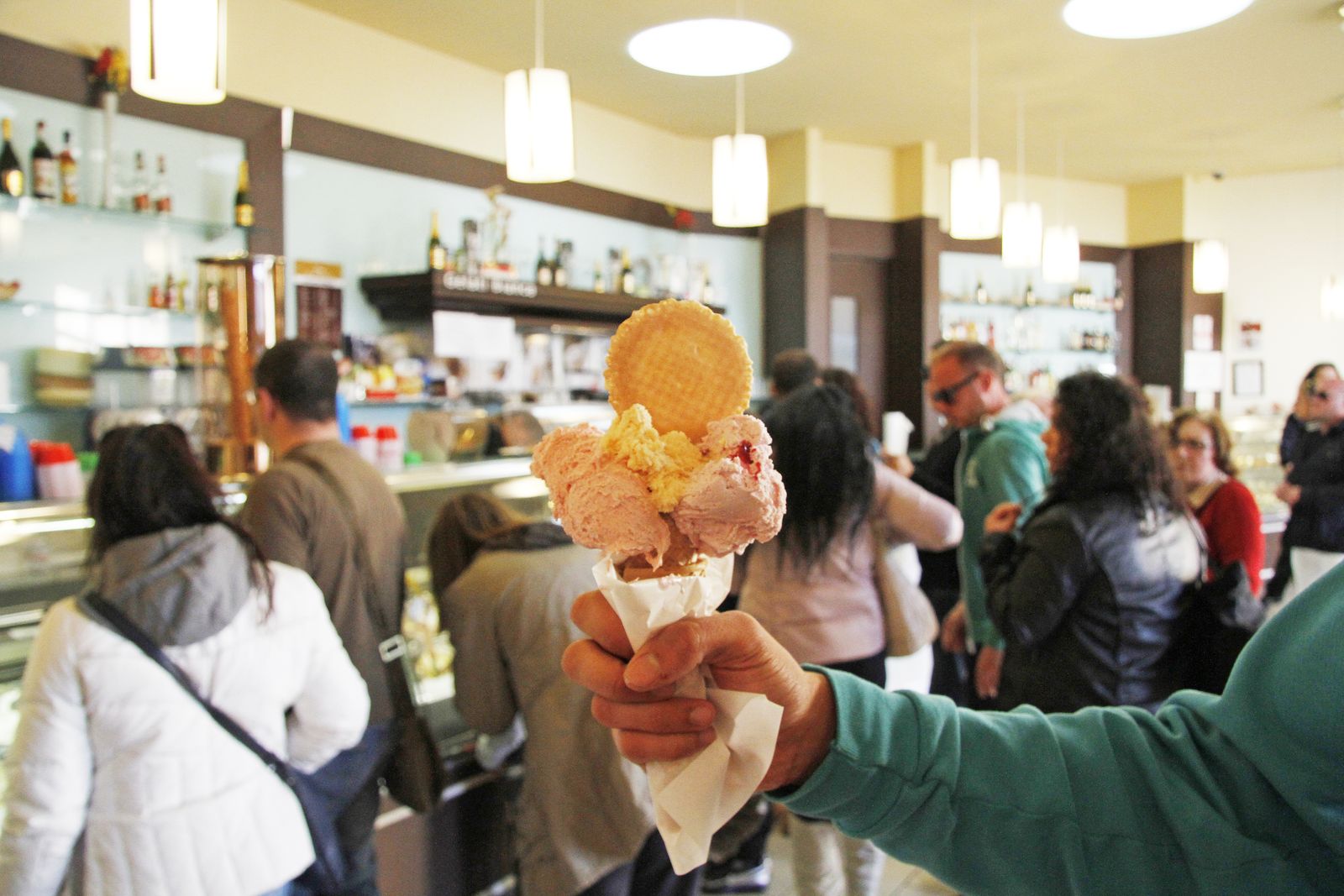 Una mano tiene un cono di gelato artigianale avvolto in un tovagliolino di carta: palline di gelato rosa con un ciuffo di crema gialla al centro e una cialda rotonda infilata in cima; sullo sfondo, sfocato, l'interno luminoso di una gelateria
