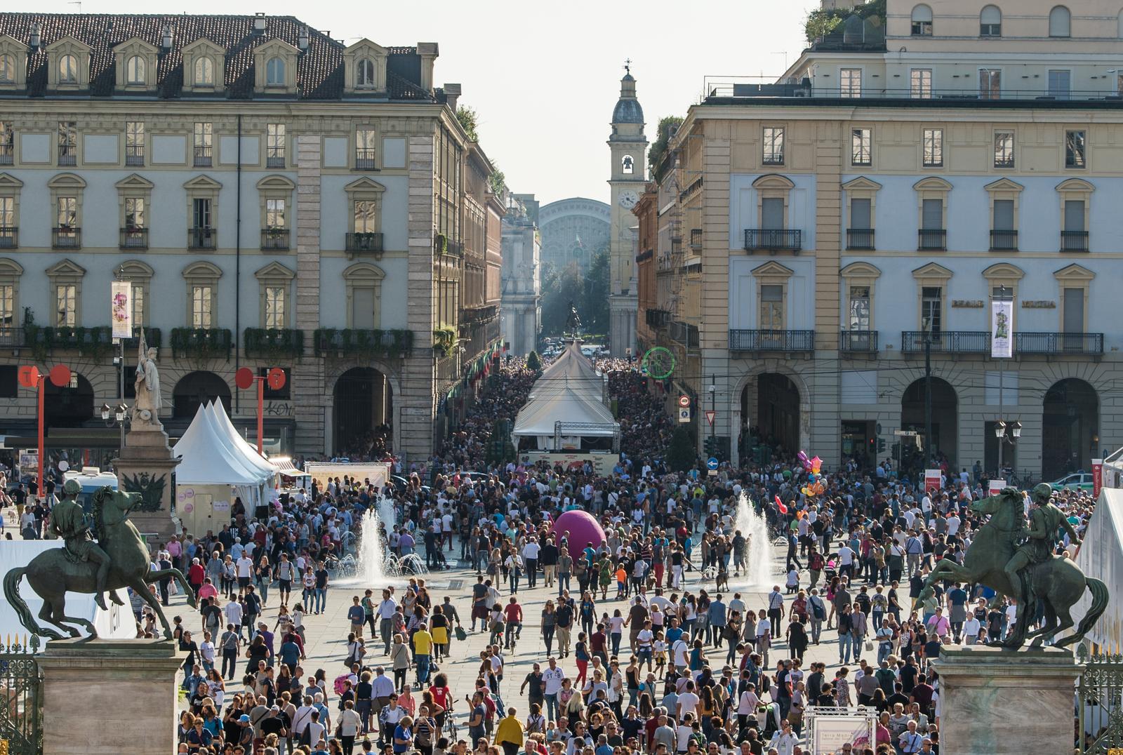 Piazza Castello a Torino in una giornata di sole, piena di gente che passeggia tra gli stand del festival: tensostrutture bianche, due fontane a getto e una grande sfera rosa; in primo piano due statue equestri in bronzo, sullo sfondo i palazzi porticati color crema e l'inizio di via Roma