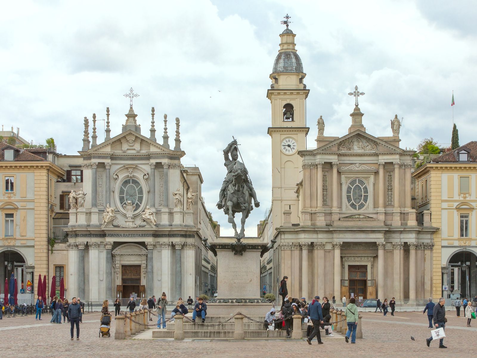 Piazza San Carlo a Torino di giorno, con il cielo nuvoloso: al centro il monumento equestre in bronzo di Emanuele Filiberto su un alto basamento di pietra, alle spalle le chiese gemelle di Santa Cristina e San Carlo, ai lati i palazzi porticati color crema e in primo piano alcuni passanti